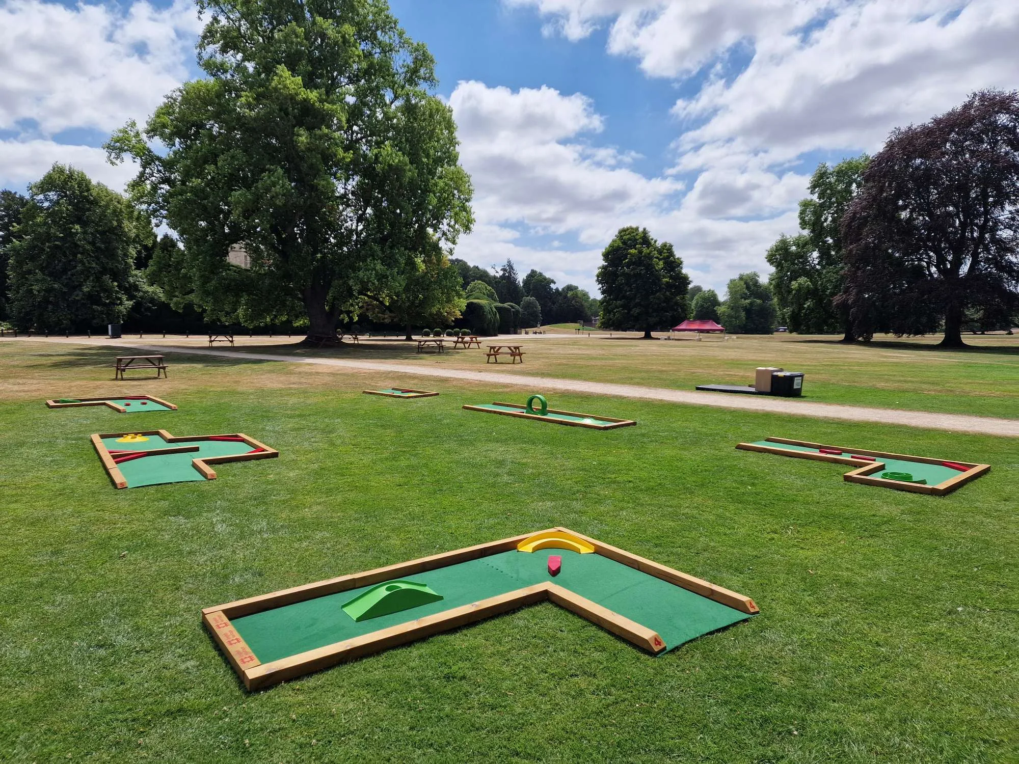 Six Putterfingers semi-permanent mini golf courses laid out on a manicured lawn at an English Heritage estate, each wooden-framed green felt hole featuring colourful obstacles including arches, ramps and bumpers, with mature trees and picnic benches visible across the sunny parkland.