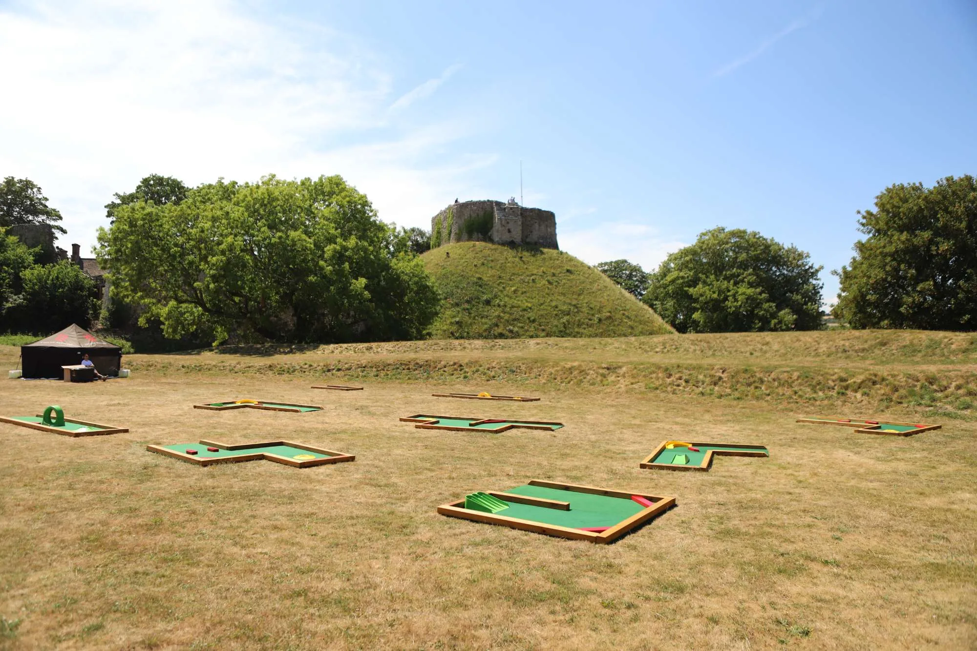 Six semi-permanent mini golf course sections with green astroturf and colourful obstacles are laid out across a dry grass field, with a grass motte and ancient stone keep ruin rising prominently behind them under a clear blue sky at an English Heritage site.
