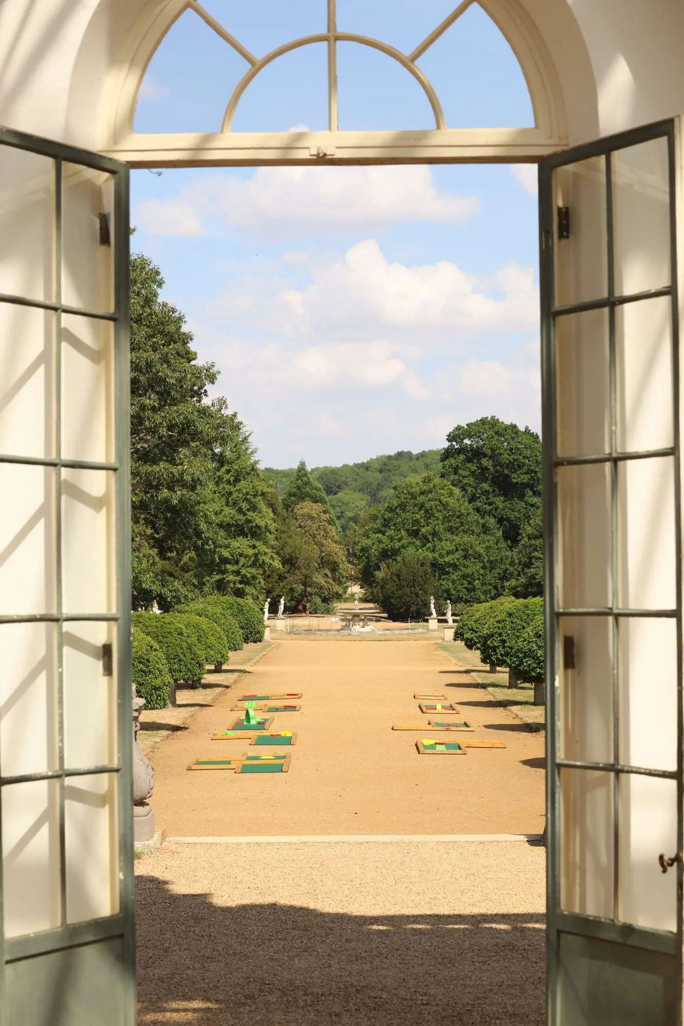 French doors open onto a formal gravel garden avenue lined with trimmed hedges and classical statues leading to a fountain, with six Putterfingers mini golf course mats laid out symmetrically on either side of the path beneath a blue summer sky.