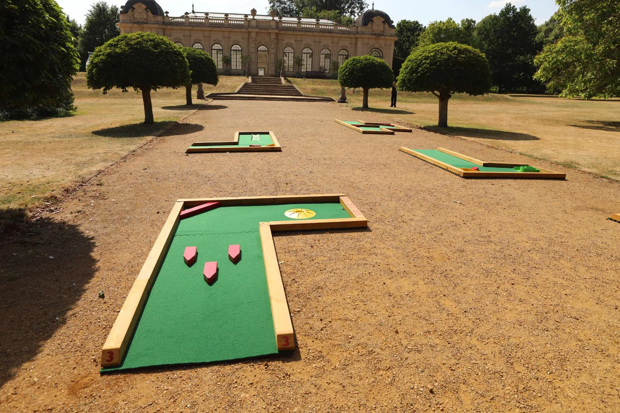 Three Putterfingers semi-permanent mini golf course holes with green felt surfaces, wooden frames, and pink arrow obstacles are laid out on a dry gravel lawn in front of an ornate neoclassical English Heritage orangery building flanked by clipped topiary trees.