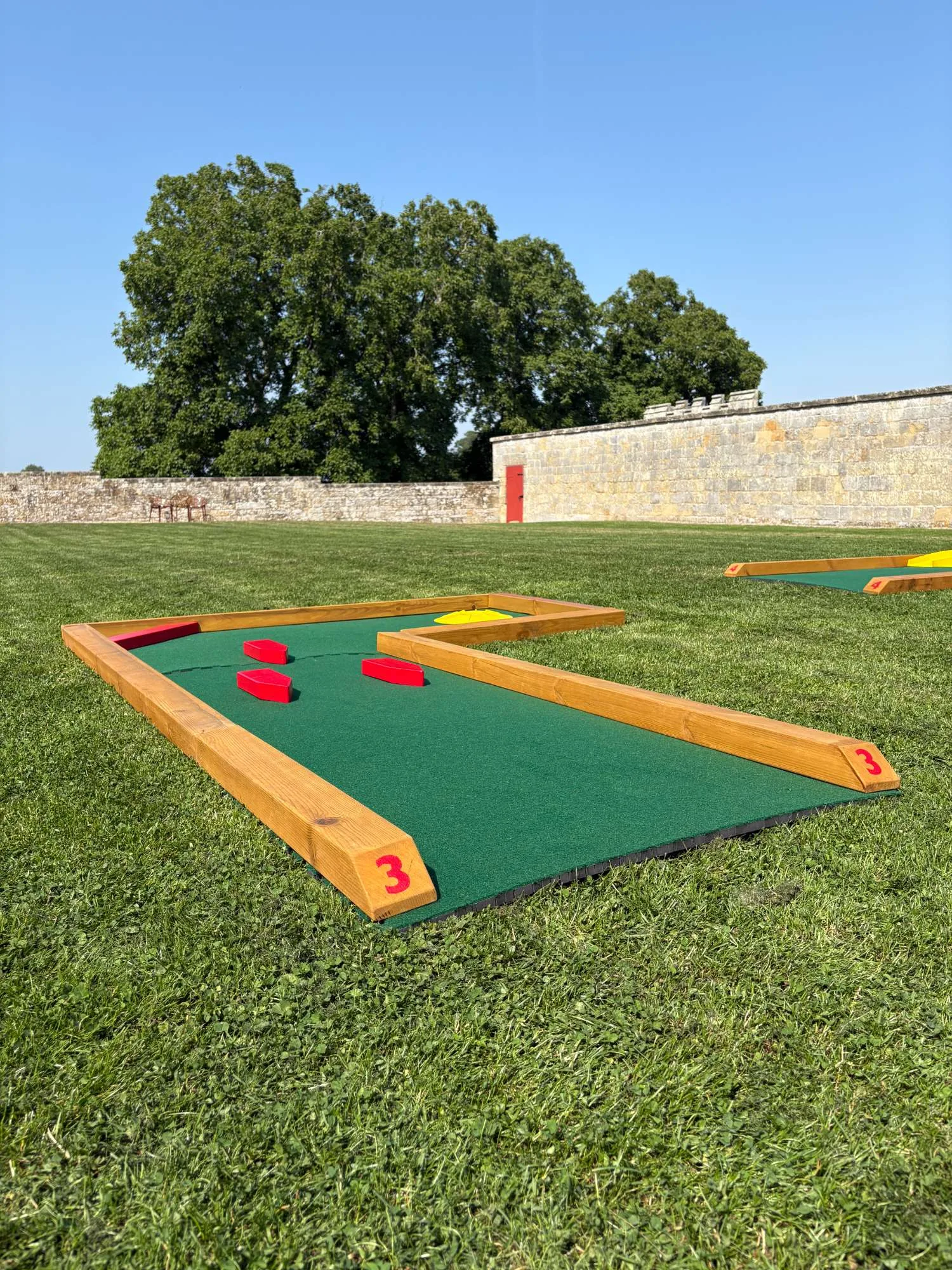 A Putterfingers semi-permanent mini golf course hole numbered 3, framed in natural timber with green artificial turf and red obstacle blocks, set on a manicured lawn beside a historic stone boundary wall at an English Heritage site on a clear sunny day.