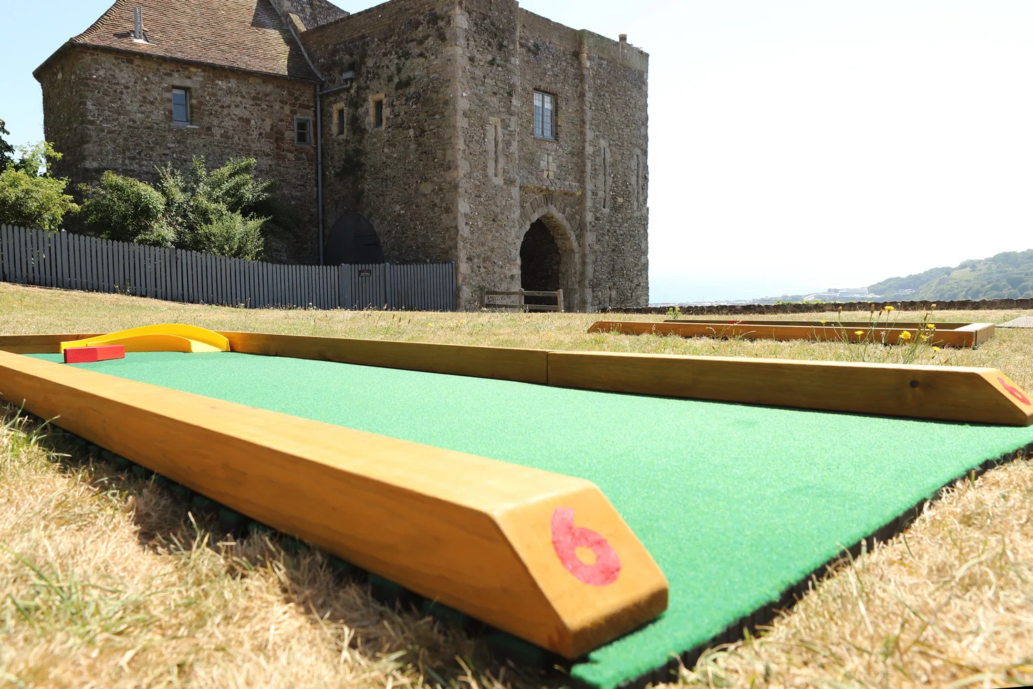 A close-up of hole 6 on a Putterfingers semi-permanent mini golf course, its green astroturf surface and wooden frame in sharp focus, with a historic English Heritage stone castle and arched gatehouse standing prominently in the background under a clear summer sky.