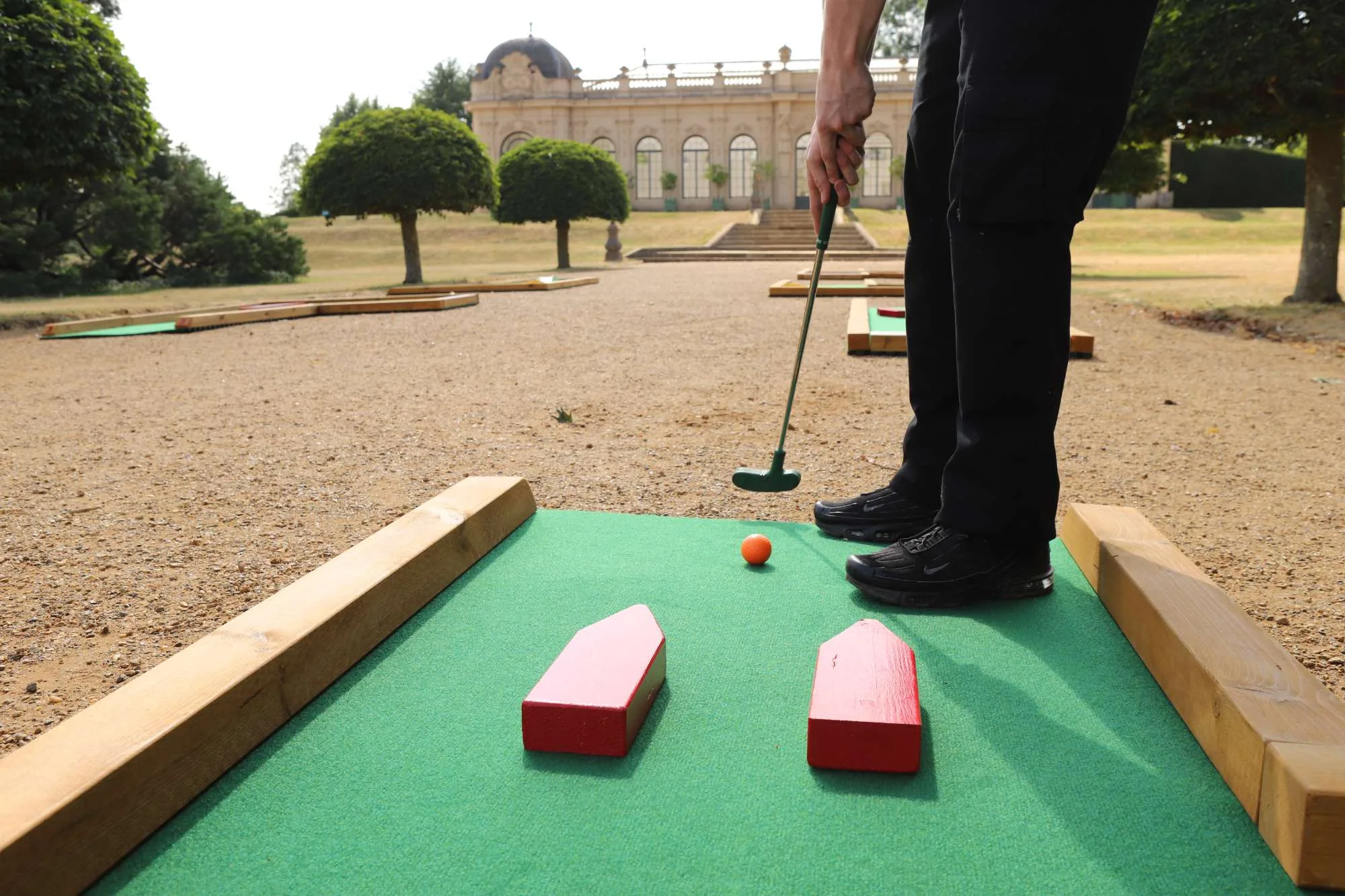 A player in black trousers and Nike trainers lines up a putt on a Putterfingers semi-permanent mini golf course set on a gravel terrace in front of a grand English Heritage orangery building, with an orange ball positioned between two red wooden obstacles on a green carpet fairway bordered by timber rails.
