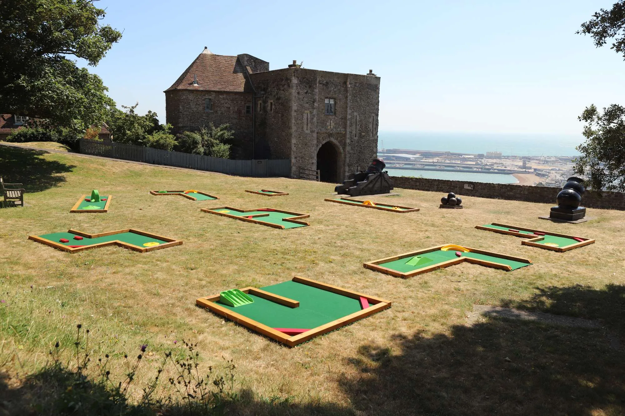 Six Putterfingers semi-permanent mini golf courses laid out on a sunny hillside lawn in front of a medieval stone gatehouse at an English Heritage site, with a harbour and coastline visible in the background and historic cannons displayed nearby.