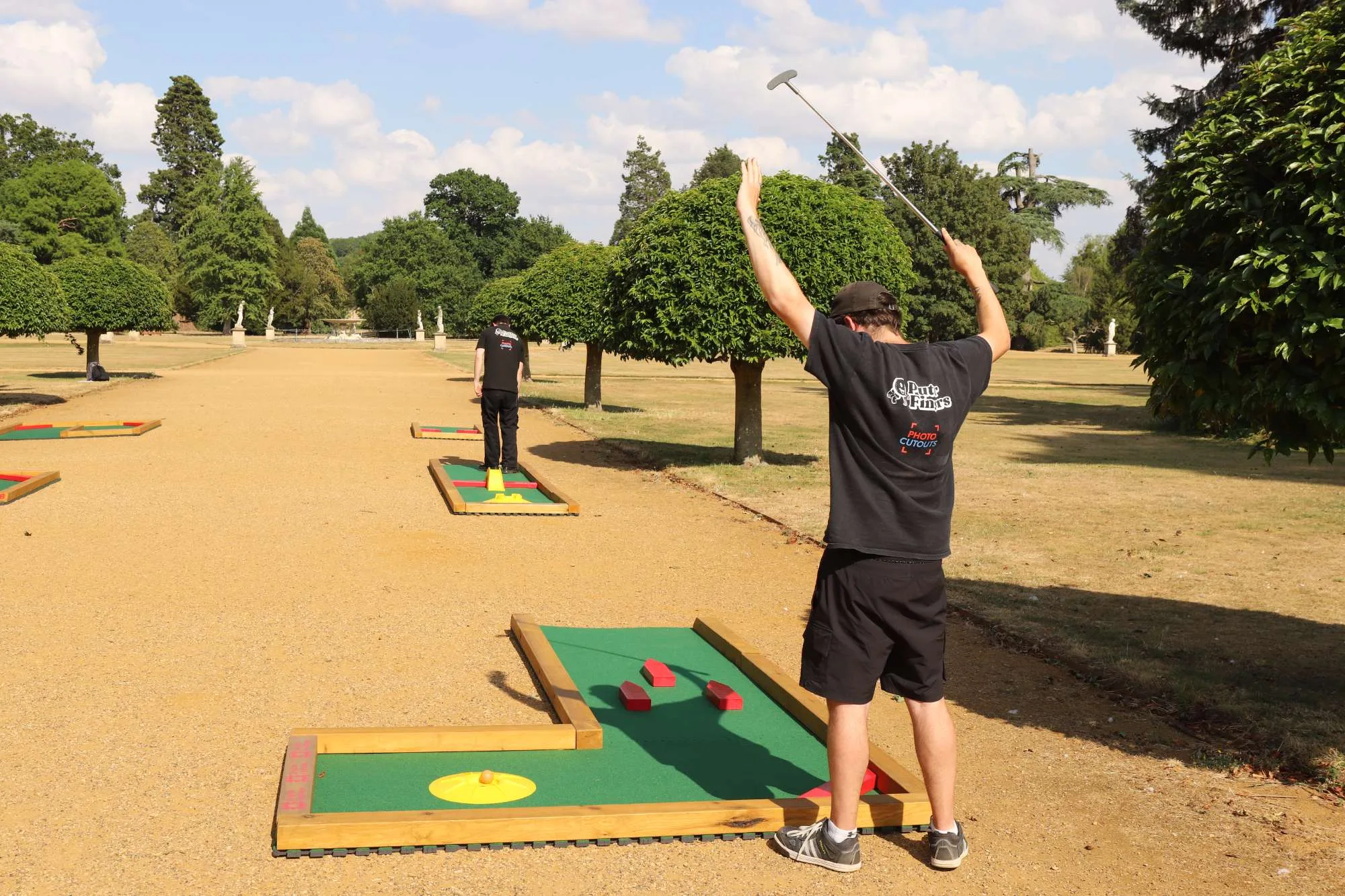 Two staff members in black Putterfingers branded t-shirts set up a semi-permanent mini golf course along a grand gravel avenue lined with manicured trees at an English Heritage estate, with one player raising a putter triumphantly above his head.