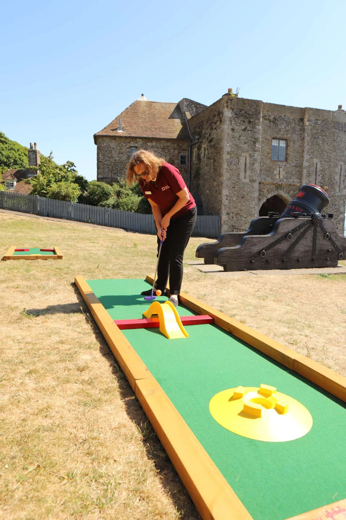 A staff member wearing an English Heritage uniform putts an orange ball on a Putterfingers semi-permanent mini golf course set up on the grounds of a historic stone castle on a sunny day, with a decorative cannon prop visible in the background.