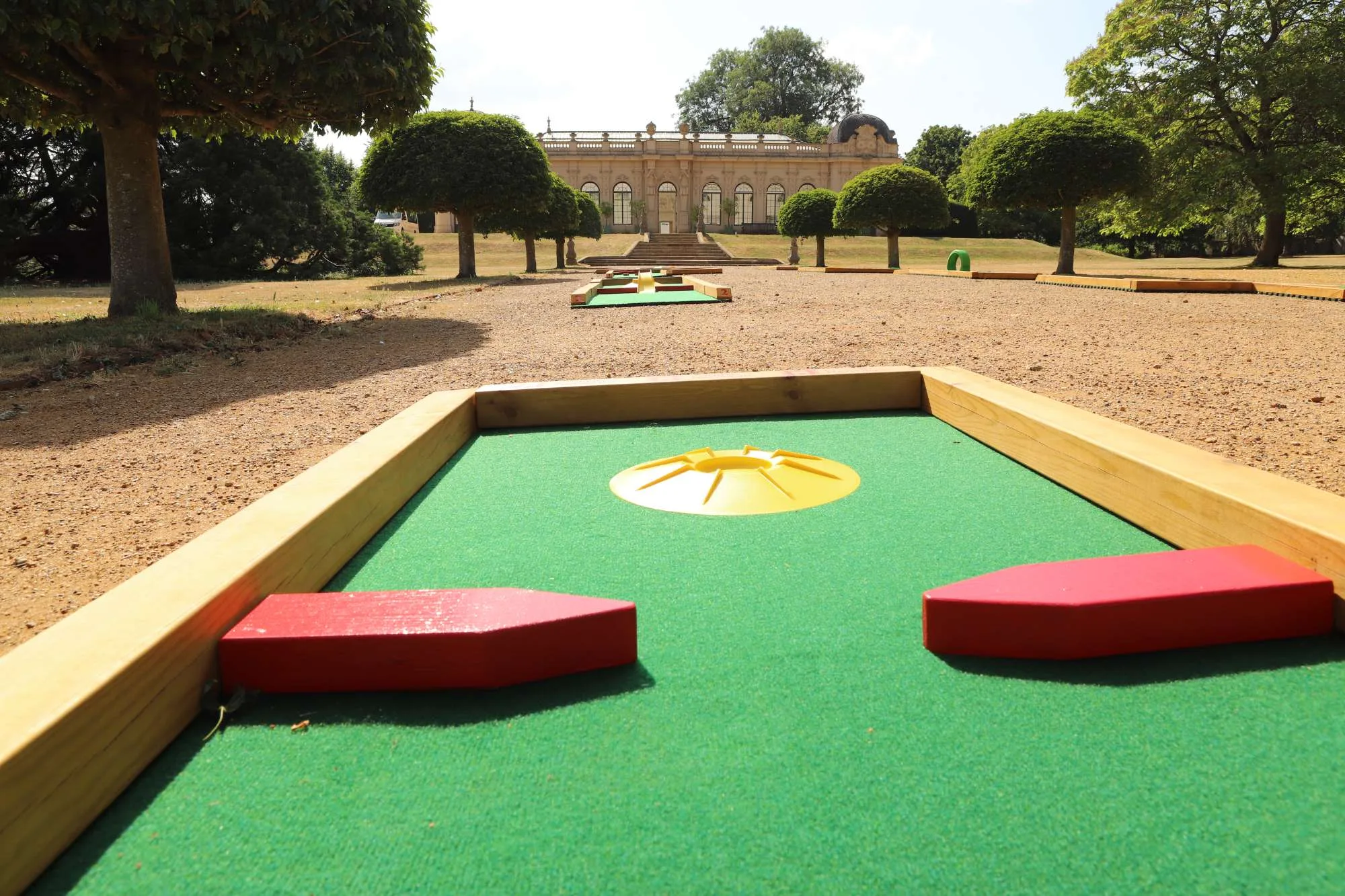 A close-up of a Putterfingers semi-permanent mini golf course hole with a green artificial turf surface, red wooden obstacles, and a yellow sun-shaped feature, set against the grand neoclassical orangery building and manicured topiary trees of an English Heritage estate.