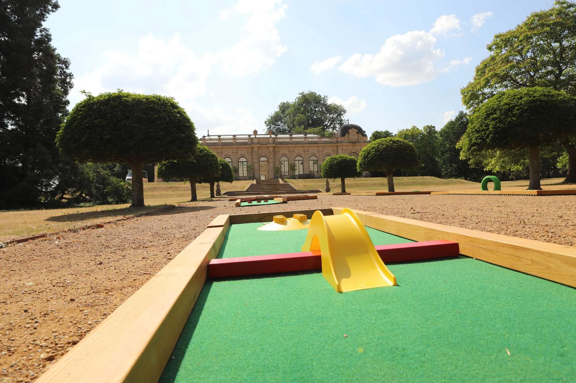 A Putterfingers semi-permanent mini golf course hole in the foreground, featuring a yellow arch obstacle and red barriers on artificial green turf, set against the grand neoclassical orangery and manicured globe-topiary trees of an English Heritage estate on a sunny day.