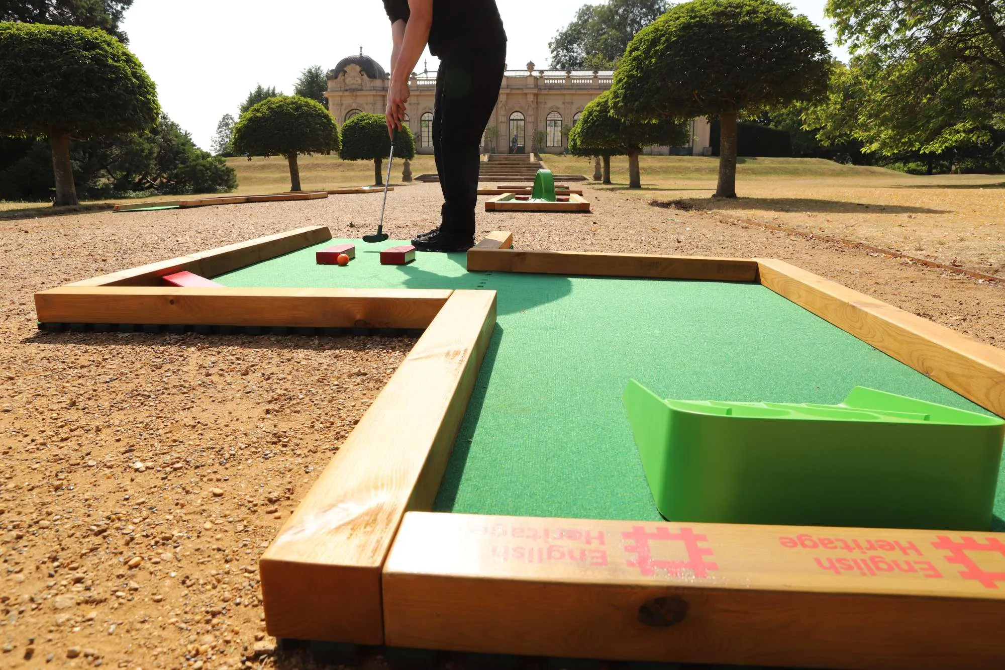 A person dressed in black putts an orange ball on a Putterfingers semi-permanent mini golf course set among gravel pathways and manicured trees, with a grand English Heritage pavilion building visible in the background.