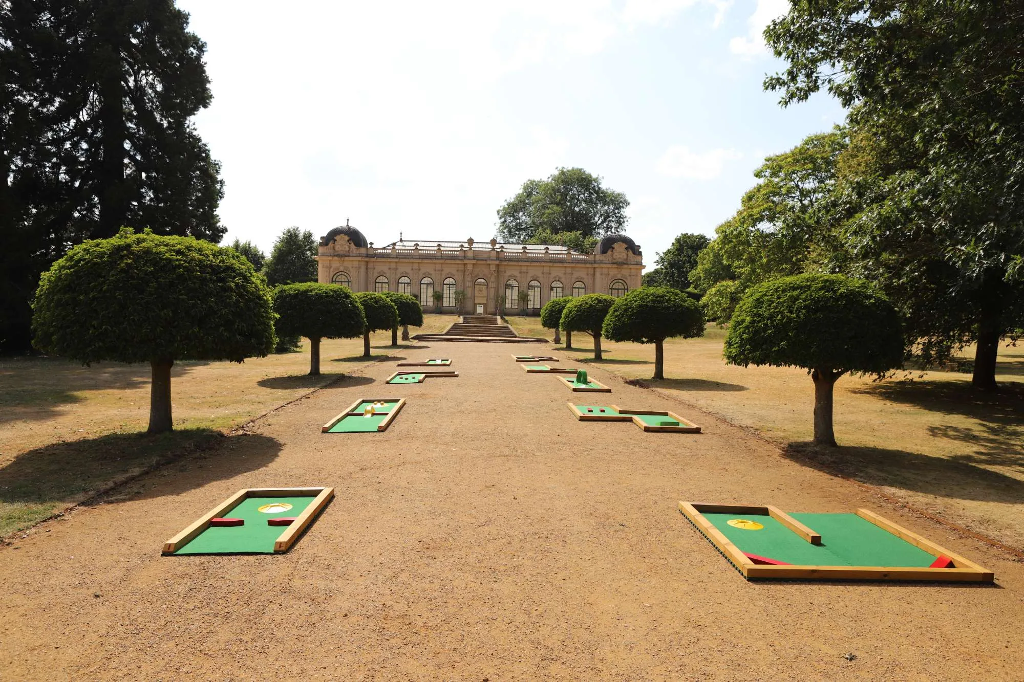 A row of Putterfingers semi-permanent mini golf courses laid out along a grand gravel avenue flanked by neatly clipped lollipop trees, leading toward an ornate Baroque orangery building at an English Heritage site on a sunny summer day.