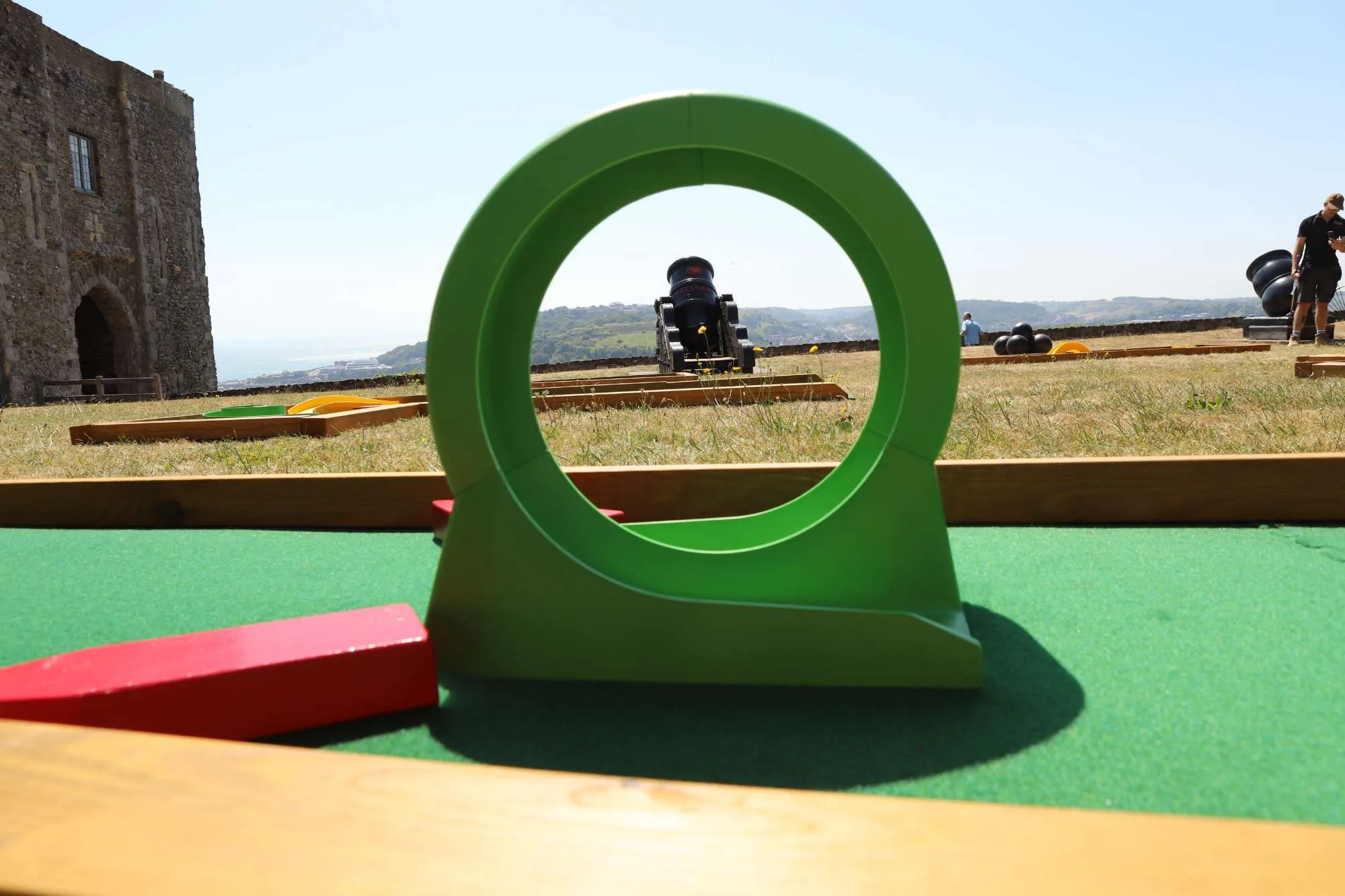 A bright green circular loop obstacle on a Putterfingers mini golf course, with a historic stone castle wall to the left and a black heritage cannon visible in the background across the manicured grounds on a sunny day.