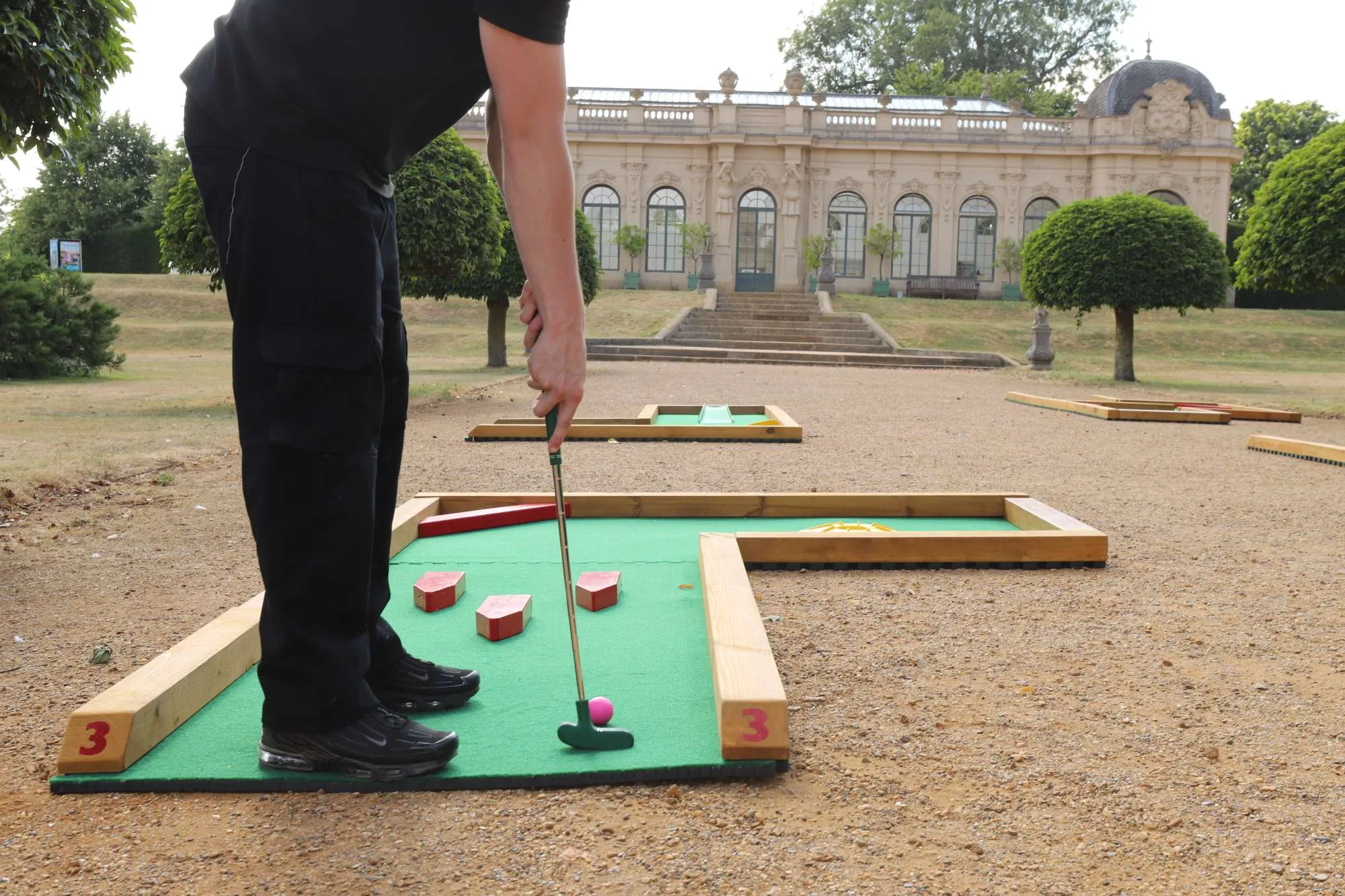 A person dressed in black leans forward to putt a pink golf ball on hole 3 of a Putterfingers semi-permanent mini golf course, set on a gravel path in front of a grand ornate English Heritage building surrounded by manicured trees.