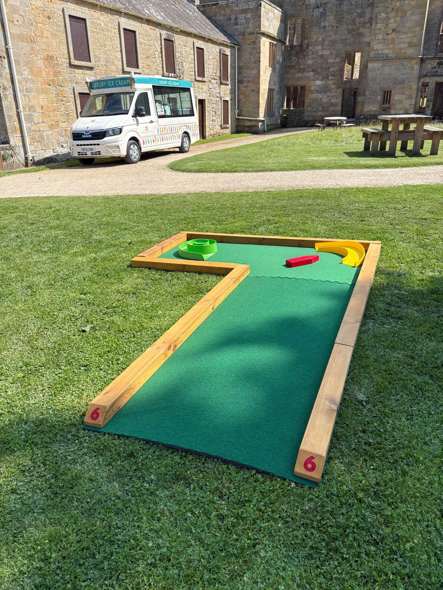 Putterfingers semi-permanent mini golf hole number 6, featuring a green astroturf surface with wooden borders, colourful obstacles including a yellow ramp and red block, set on a grass lawn in front of a historic English Heritage stone building with a luxury ice cream van parked nearby.