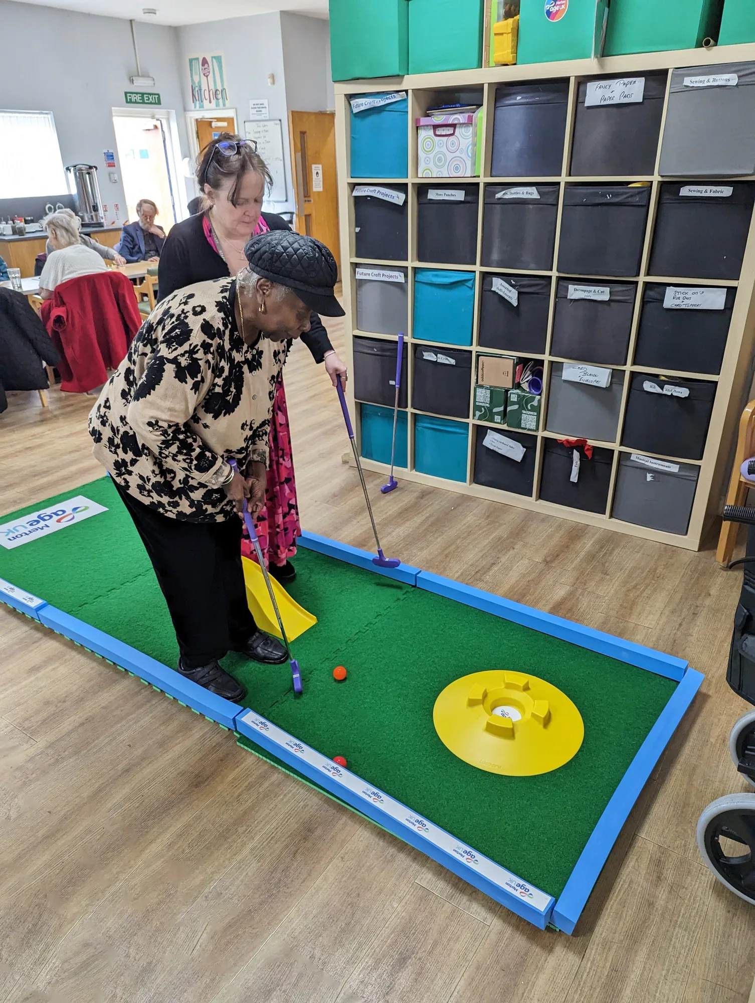 An elderly woman bends forward to putt an orange ball on a Putterfingers indoor mini golf green with a yellow target hole, assisted by a carer in a black cardigan at an Age UK centre with craft storage shelving visible in the background.