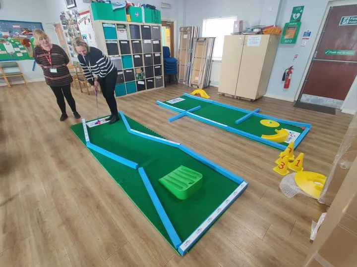 Two women enjoy indoor crazy golf on a Putterfingers portable mini golf course set up inside an Age UK care home activity room, one preparing to putt while the other watches.
