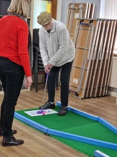 An elderly resident wearing a flat cap putts a golf ball on a Putterfingers portable mini golf green indoors, while a staff member in a red jumper looks on.