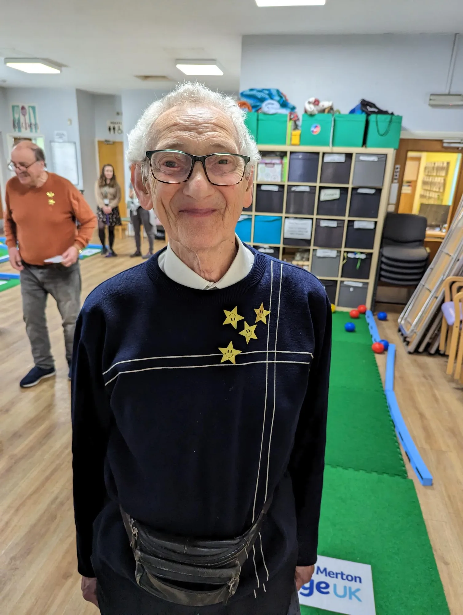 An elderly man wearing a navy sweatshirt decorated with gold star stickers and a bum bag smiles at the camera during a Putterfingers crazy golf session at an Age UK event, with a putting green mat and storage shelving visible behind him.