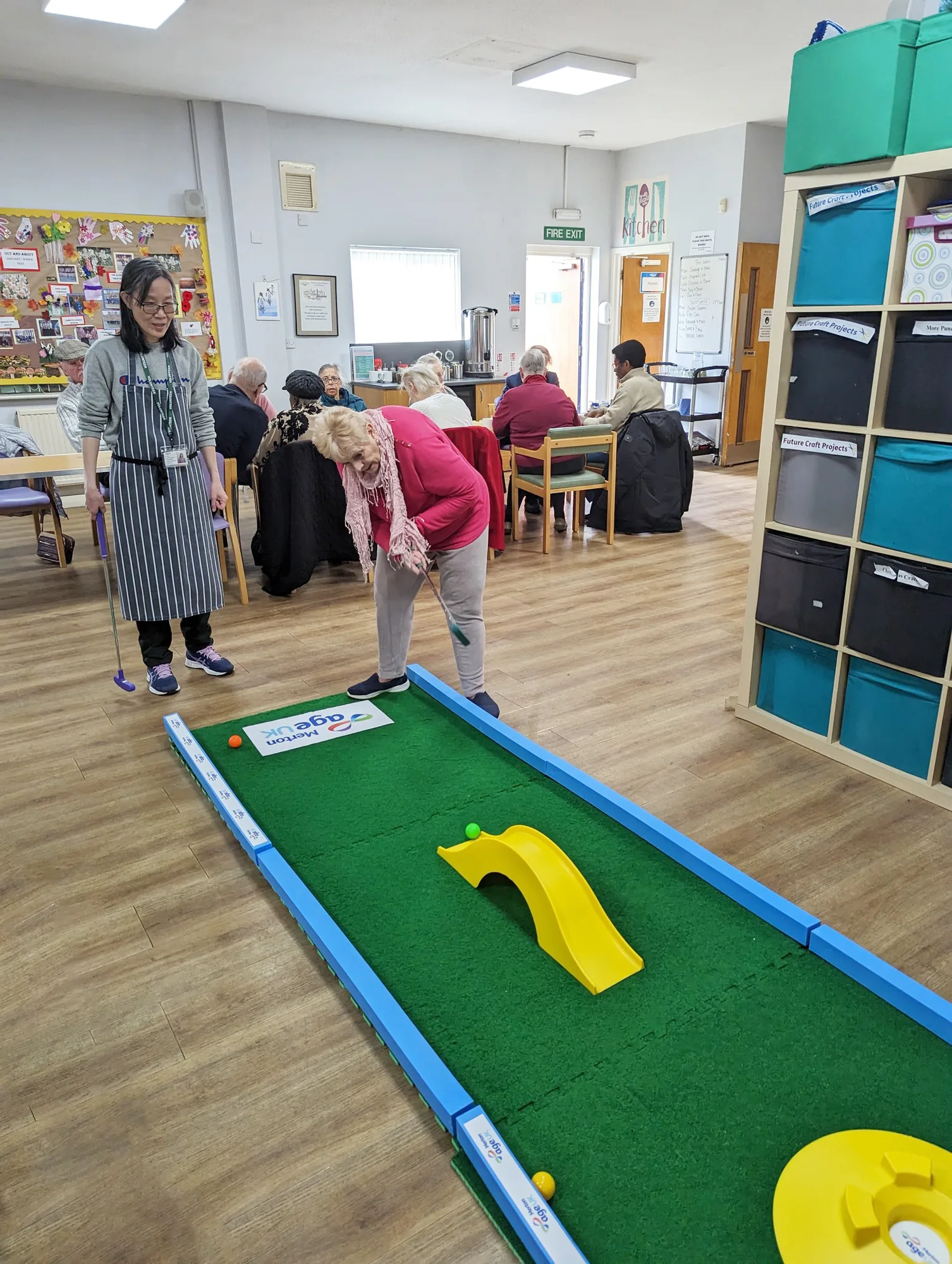 An elderly woman in a pink cardigan putts on a Putterfingers Age UK branded indoor crazy golf course while a care worker in a striped apron looks on, with other seniors socialising at tables in the background of a bright community room.