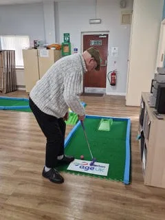 An older adult putts a golf ball on a Putterfingers indoor mini putting green set up inside an Age UK care home activity room, with a fire extinguisher and wooden flooring visible in the background.