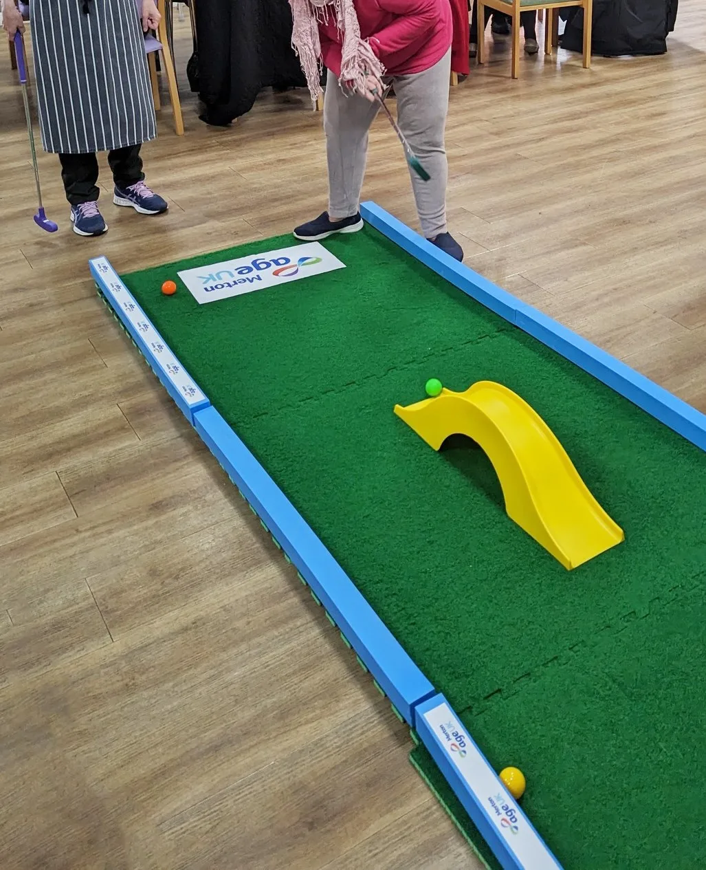 A person in a pink top and grey trousers putts a golf ball along a Putterfingers indoor crazy golf putting green branded with the Age UK Merton logo, featuring a yellow bridge obstacle and coloured balls on artificial grass.